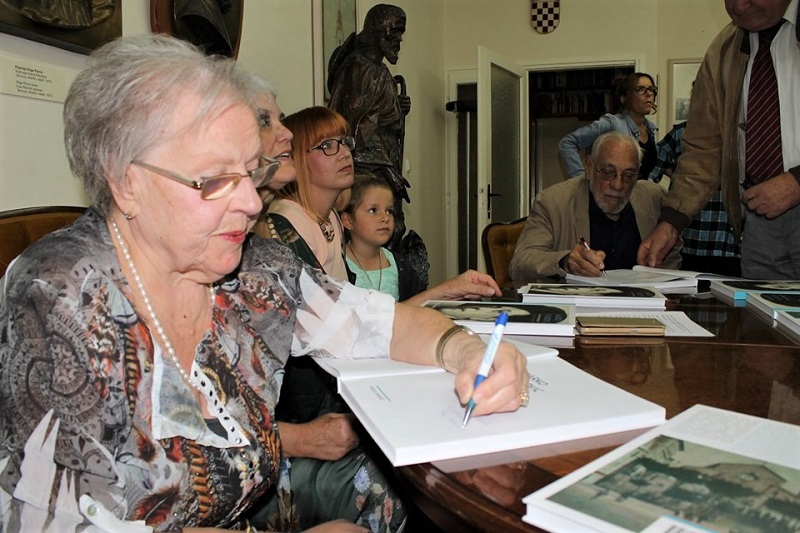 Marija Barbieri na promociji monografije o Marku Vuškoviću u Galeriji <em>Ivan Rendić</em> u knjižnici u Supetru, autorica i umjetnikov unuk upisuju posvete u knjigu